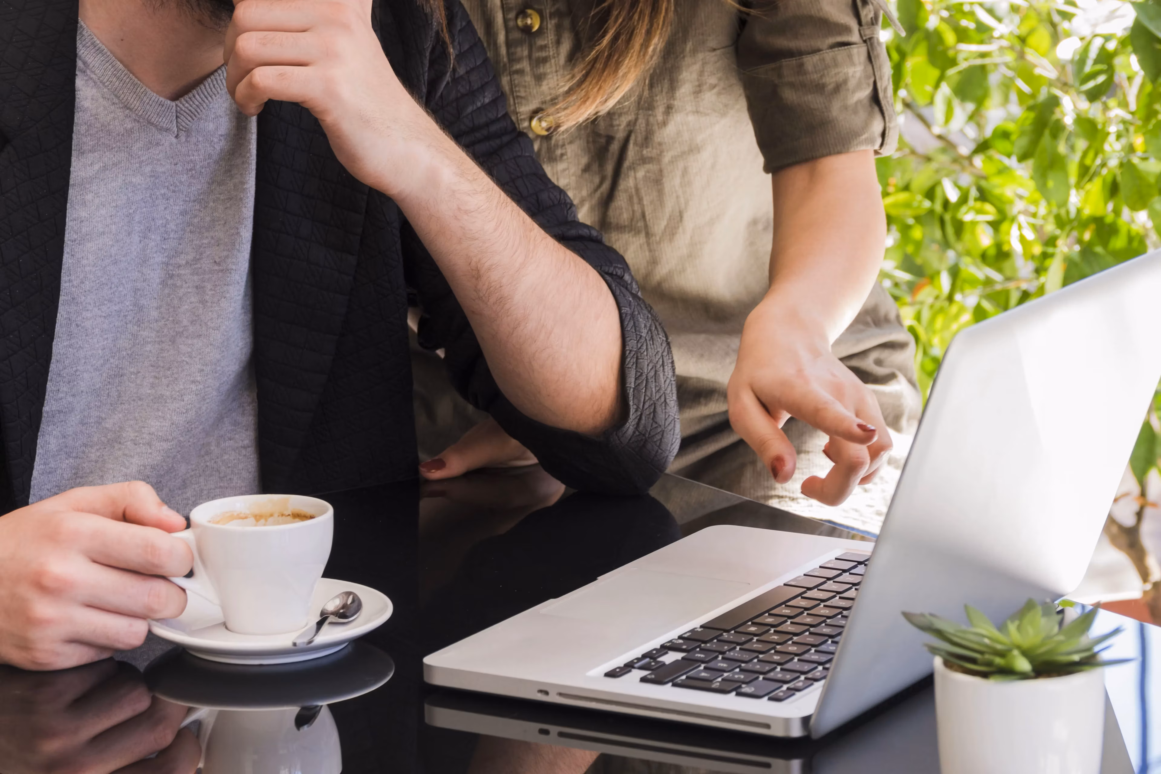 Two coworkers at a shared desk, one pointing at a laptop screen while discussing a project
