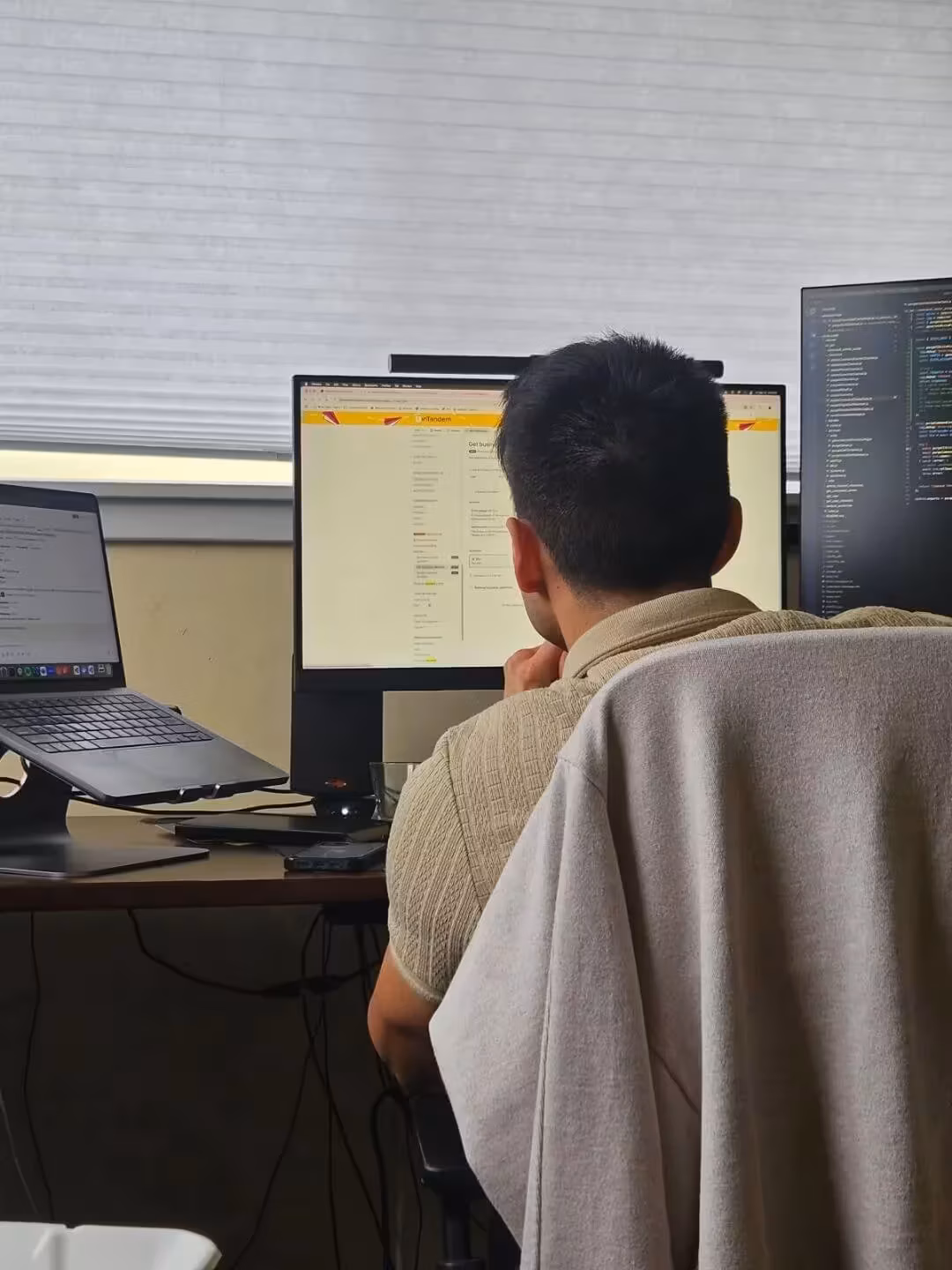 Man seated at a workstation with three monitors, reviewing technical documentation and programming code